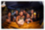 Six women in black and gold clothing raise drinks at a lively birthday party, smiling by a wood bar; Legend poster in background.