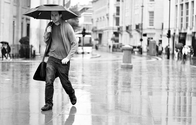 Man walking with umbrella on rainy city street. Black and white image with wet pavement, buildings in background, and somber mood.