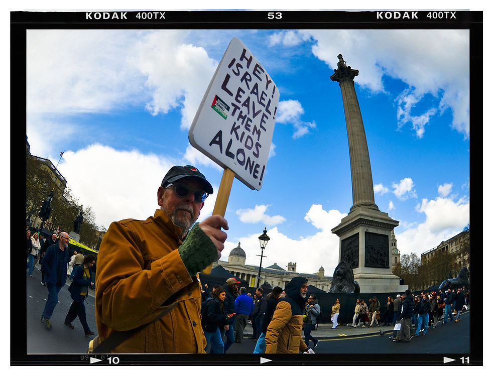 Protester holding a sign reading "Hey! Israel! Leave Them Kids Alone!" in a busy square with a tall column. Sky is partly cloudy. Together Alliance, London, March 2026