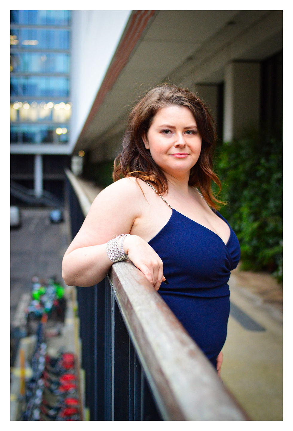 Woman in a blue dress leans on a railing outdoors, wearing a sparkling bracelet. Background features a building and greenery.