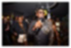 Stephen Rowe speaks into microphone at a party, with a crowd and black-and-gold balloons behind him.