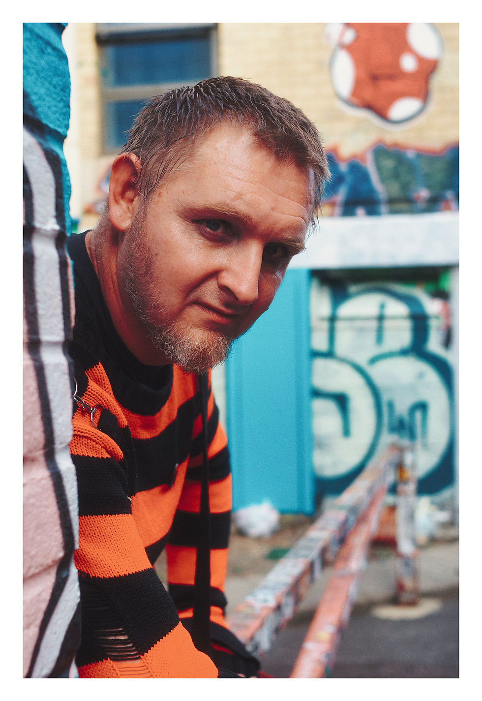 Close-up of a man in an orange-and-black striped sweater leaning forward against a graffiti-covered wall, looking serious.