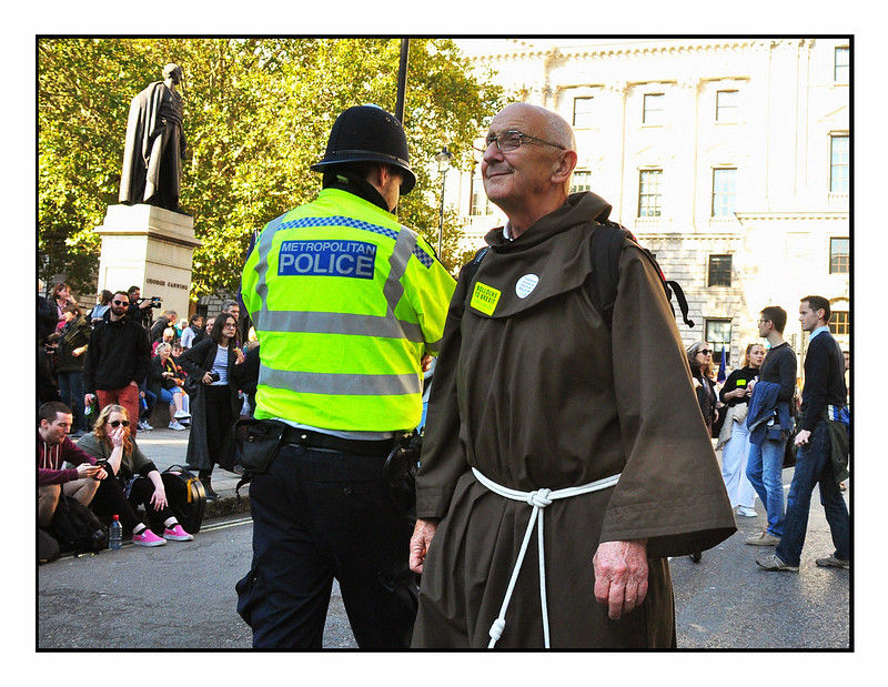 A man in a monk robe and a police officer in a high-visibility jacket stand in a lively street. Sunlight filters through trees.