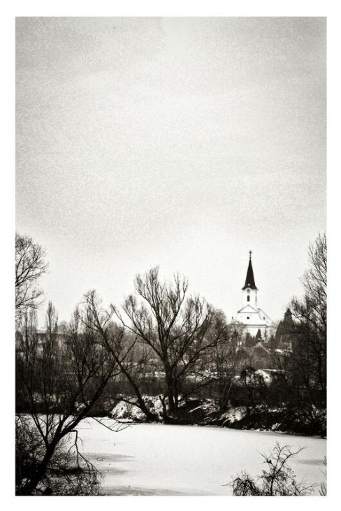 A black and white image of a distant church with a tall steeple, surrounded by bare trees. A frozen river is in the foreground, creating a serene winter scene. Soporna, Slovakia
