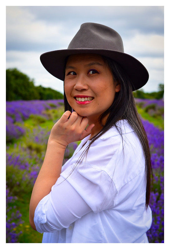 Woman in a white shirt and hat smiles in a lavender field. Purple flowers and cloudy sky create a serene, joyful atmosphere.
