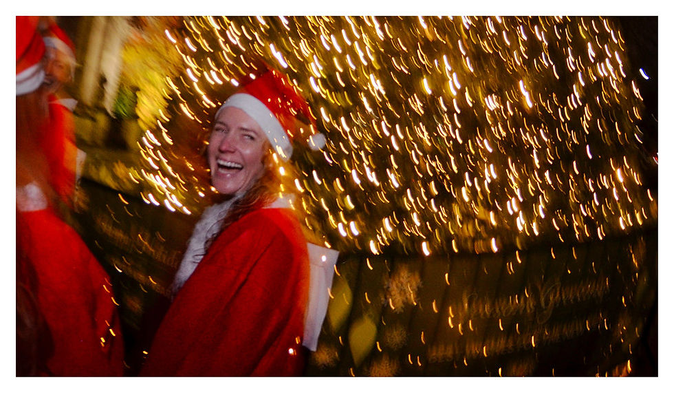 Person in Santa outfit laughing, standing near a brightly lit Christmas tree. Warm, festive atmosphere with blurred golden lights. Santa in the City Walk and Run, London