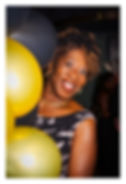 Smiling woman peeks from behind black and yellow balloons at a party, with a dark background and a joyful mood.
