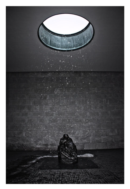 Dark statue sits under circular skylight in gray room. Rain falls through the opening, creating a somber, reflective mood. memorial to the victims of war and tyranny, Berlin