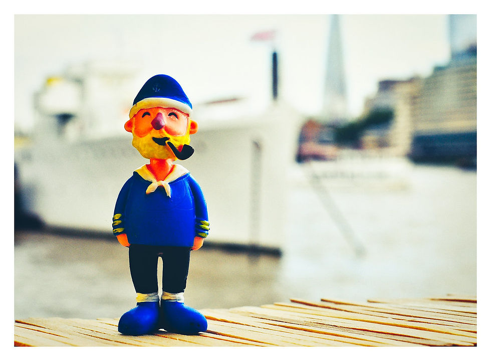 Clay sailor figurine with a pipe stands on a wooden surface. Background shows blurred boat and cityscape, creating a whimsical, nautical scene.