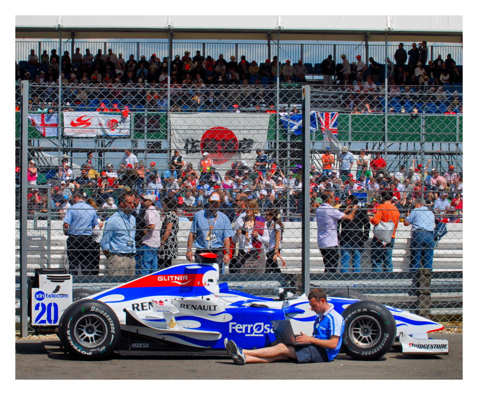 Race car with blue and white livery, man using a laptop beside it. Crowd behind a fence, flags, and banners visible. Sunny day.
