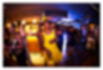 People dancing at a lively nightclub, with a woman in a bright yellow dress in front under warm and blue lights.