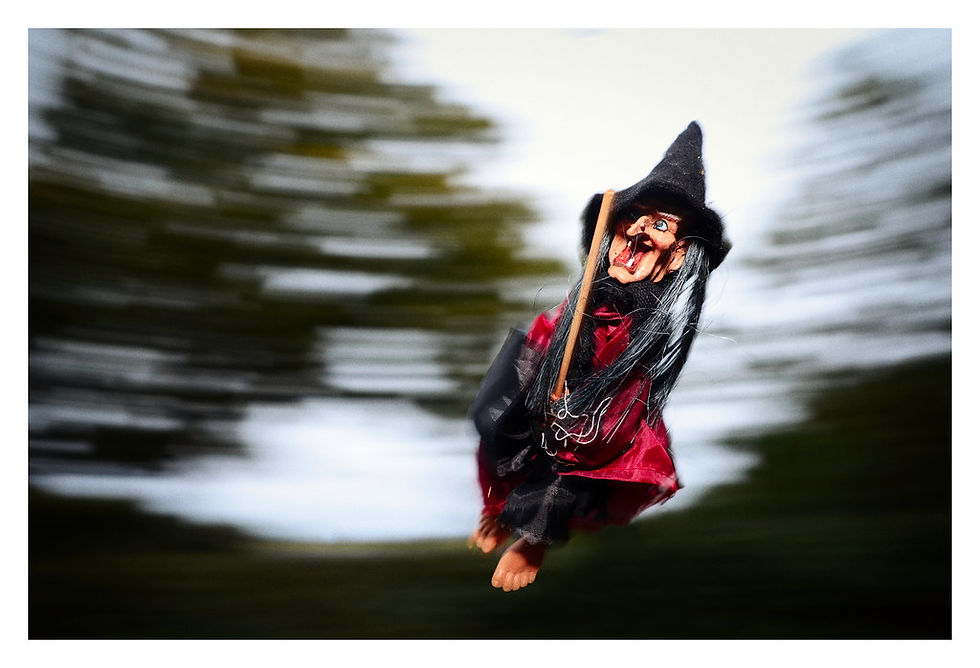 A witch doll in red and black rides a broomstick, set against a blurred green and white background, creating a sense of motion.