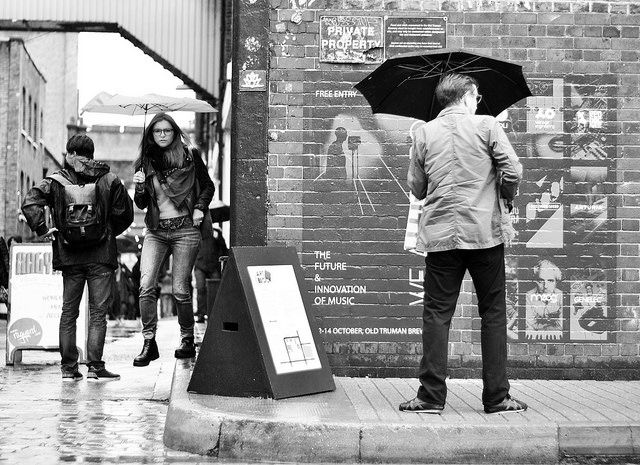 People with umbrellas walk on a rainy city street. A man reads posters on a brick wall. Monochrome setting, wet pavement, casual mood.