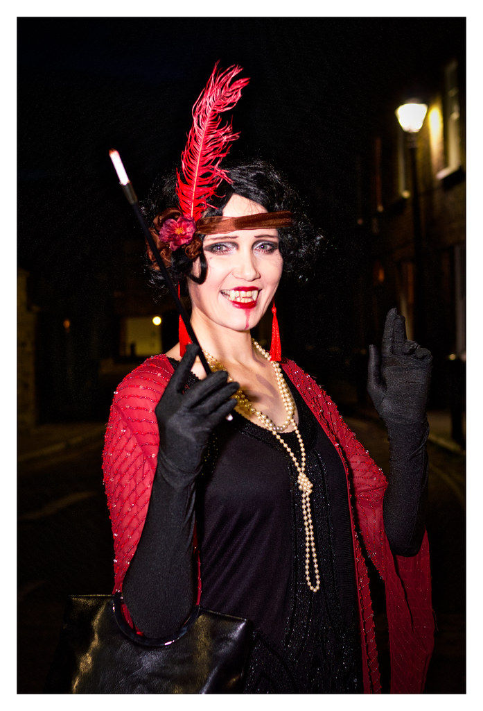 Smiling woman in 1920s flapper costume with red feather headpiece and pearls, posing on a dark street at night.