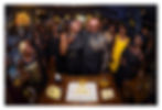 Crowded birthday party with smiling guests around a cake, black-and-gold decor, and two people at the center.