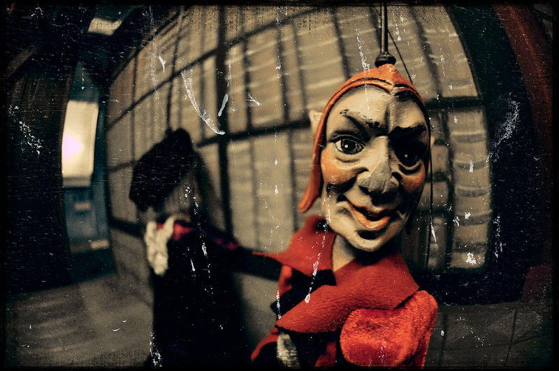 Close-up of a marionette with a mischievous grin in a red outfit. Blurred background with wooden textures adds a vintage feel.