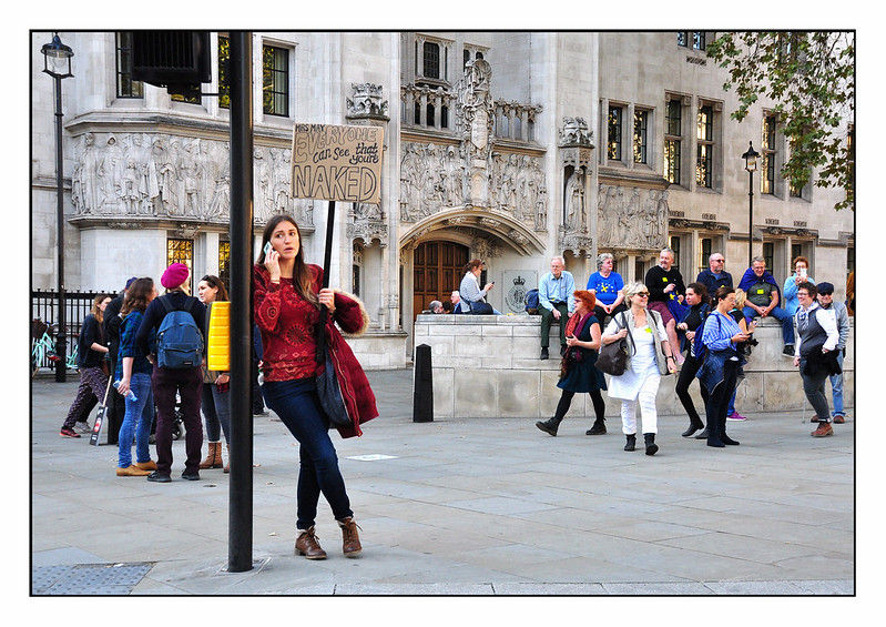 Woman on phone leans against pole with sign reading "Everyone can see you're naked" in front of historic building, surrounded by a crowd.