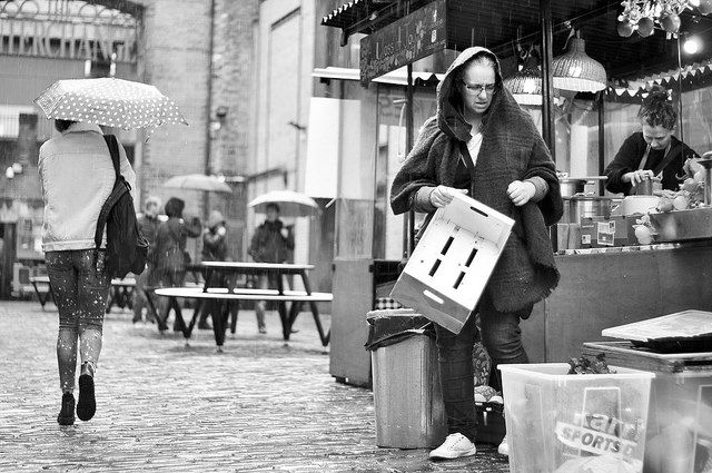 A street scene in the rain shows a person holding a box near a market stall. Another person with an umbrella walks away. Gloomy mood.