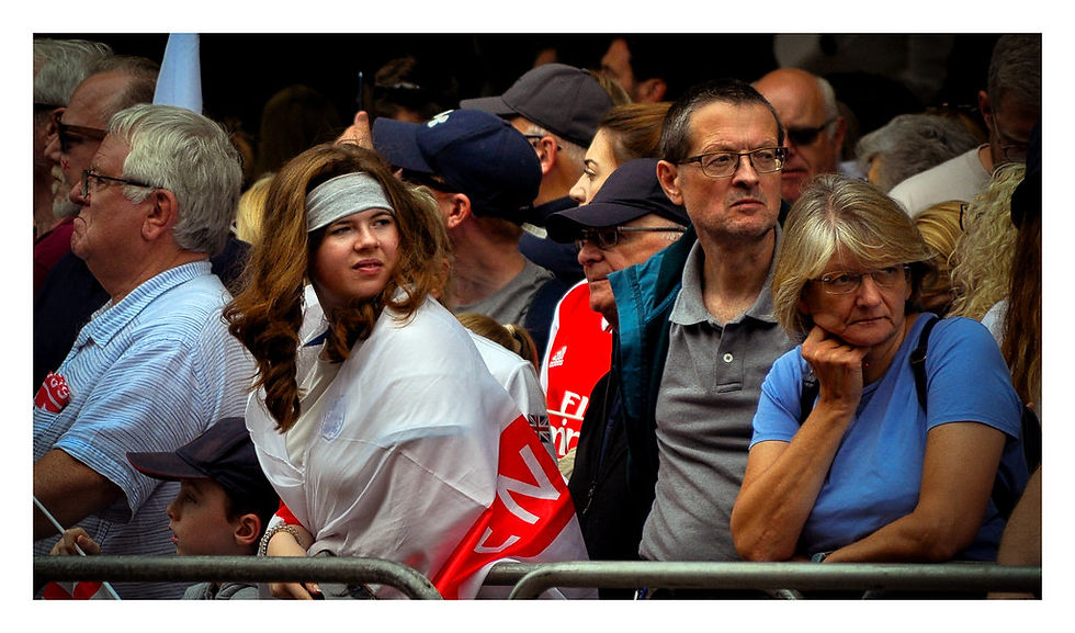 Crowd watching an event. A woman in white with a red flag, next to people looking attentive. Background filled with spectators, mixed emotions. Celebrations of England Women's Euro 2025 win at the Victory Parade