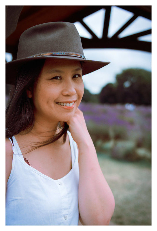 Woman in a white dress and hat smiles, standing outdoors with purple flowers and trees in the blurry background under a gazebo.
