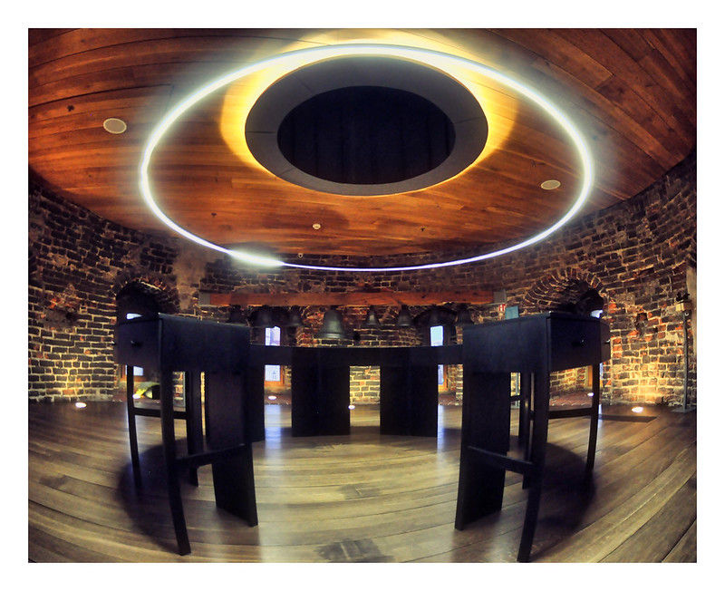 Circular room with brick walls, wooden floor, and ceiling. A glowing ring light hangs above dark wooden tables, creating a modern, serene vibe.
Desk with a circle light above it inside the Vilnius Cathedral Square Bell Tower
