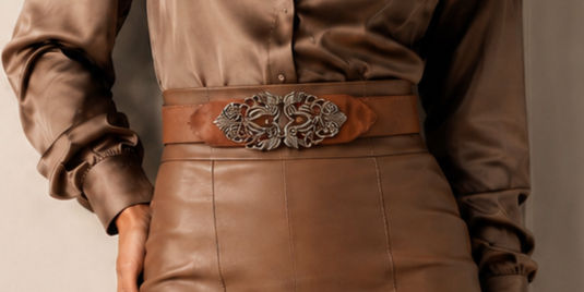 Close-up of a woman in a brown satin blouse and leather skirt, wearing an ornate belt buckle against a neutral background.