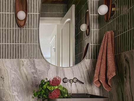 Green tile upscale bathroom. Wood and brass touches. Flowers in the sink. Brown hand towel.