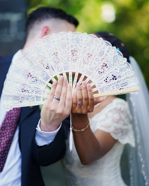 Bride and groom holding decorative fans in front of faces during portrait.