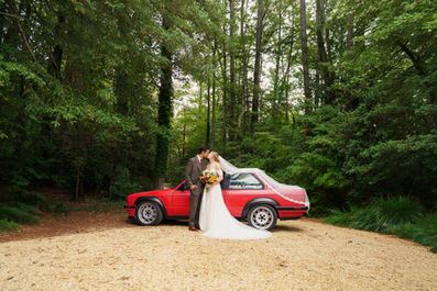 Bride and groom standing next to a vintage red car in a wooded area.