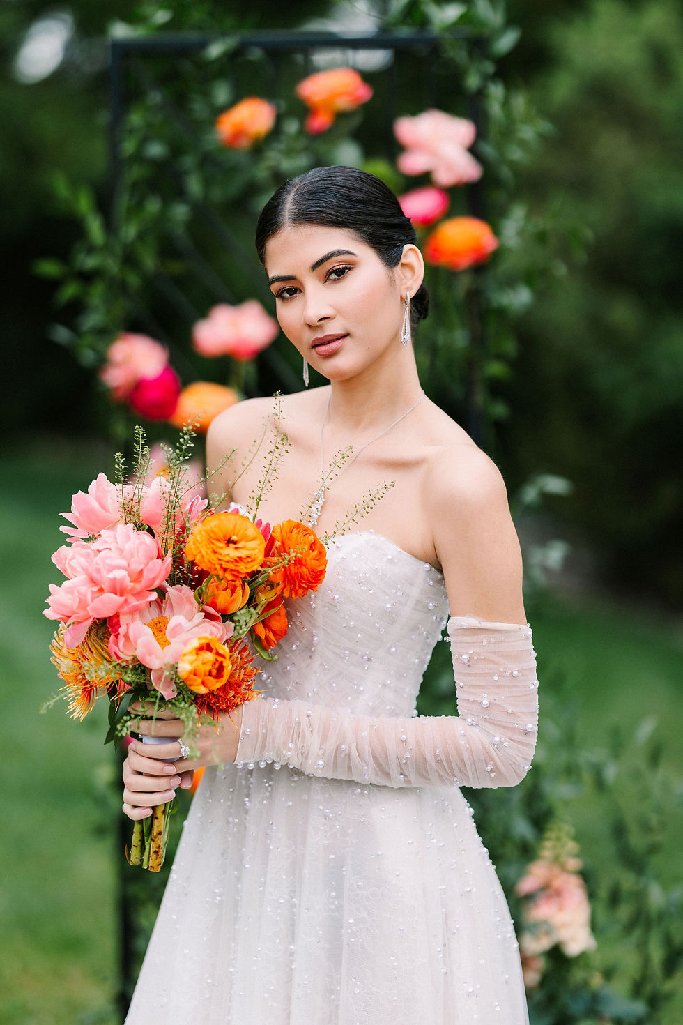 Woman in white dress holds bouquet of pink and orange flowers, standing in garden with floral arch. Elegant and serene mood.