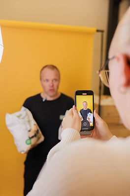 Nainen ottaa puhelimella videota miehestä keltaisen taustan edessä.