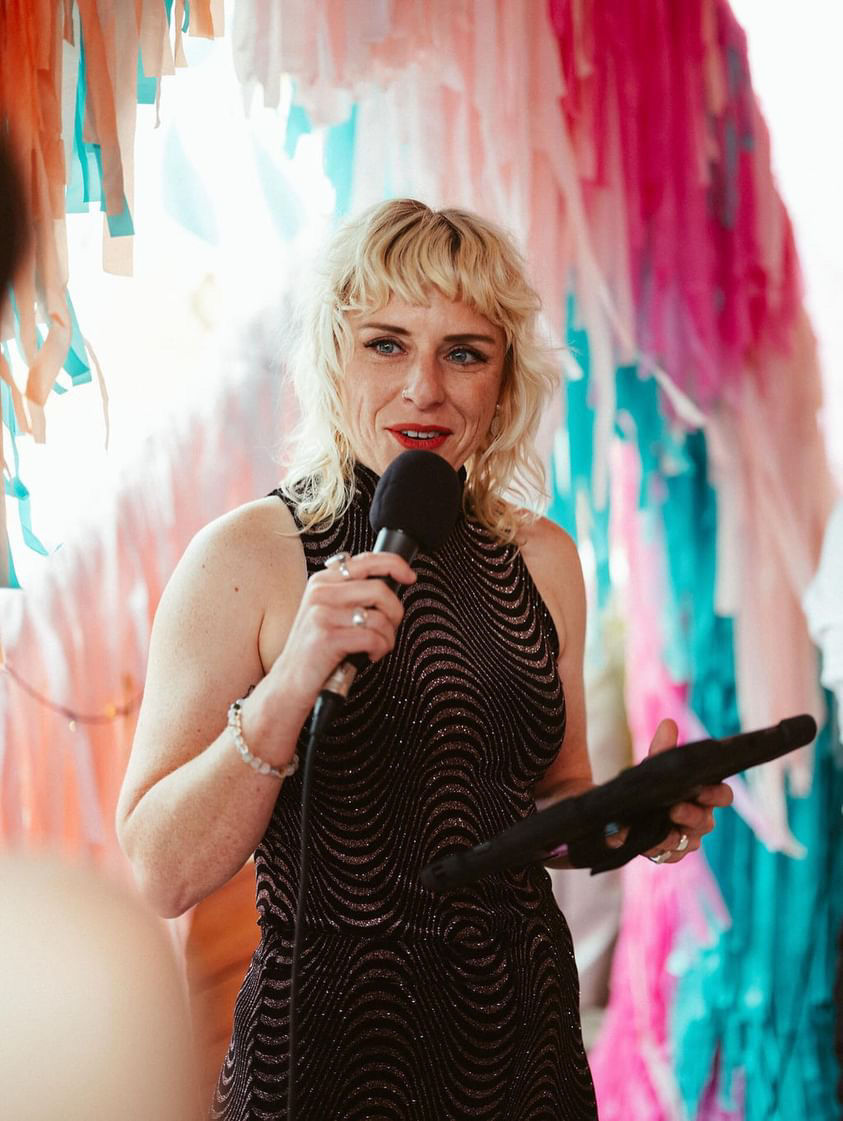 Renee the celebrant and MC has blonde wavy hair, is holding a microphone and talking into it with rainbow streamers in the background. She's smiling while she talks