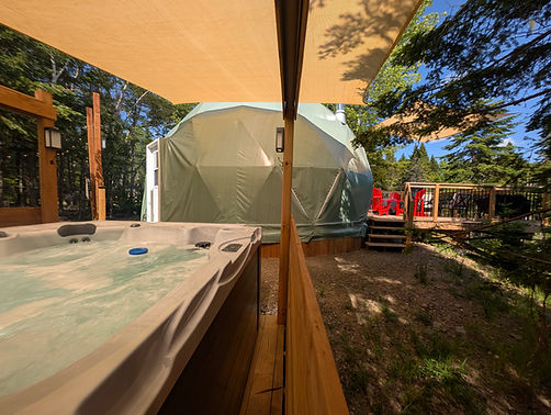 Hot tub next to a green dome in a forest, lots of privacy, there is also a deck on the front of the dome. The deck ahs red chairs, a blackstone griddle and bbq and oversized games for guests to play. This dome is at Forest Lane Domes and Experiences in Bloomfield New Brunswick