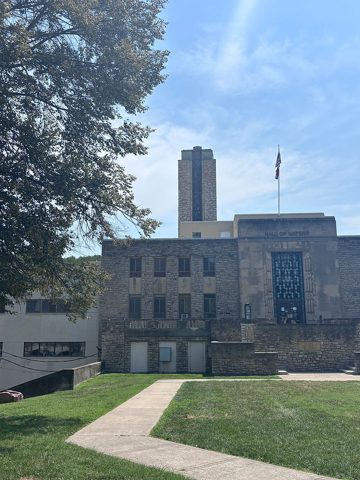 The stone facade and tower of the Hall of Waters, framed by a tree and a path across the lawn.