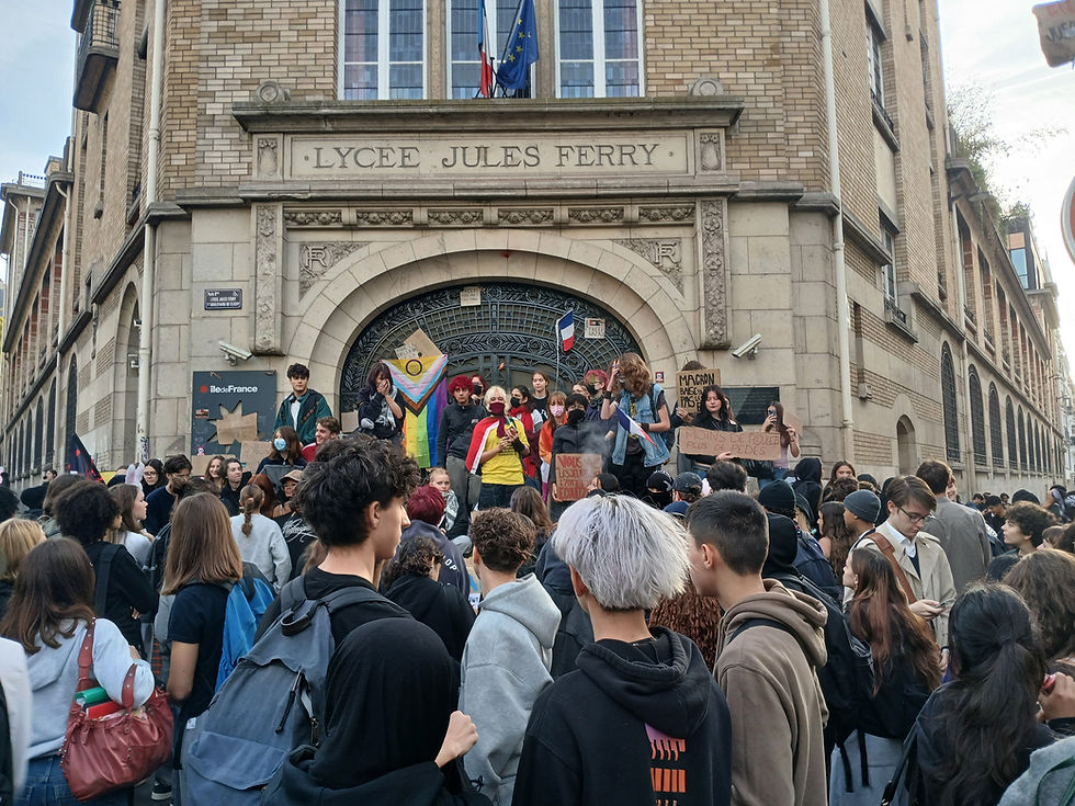 Manifestazione studentesca davanti all'ingresso del Lycée Jules Ferry a Parigi. Numerosi giovani protestano sui gradini e in strada esponendo cartelli e bandiere, tra cui la bandiera LGBTQ+.
