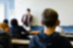 Zoom su un ragazzino seduto tra i banchi di scuola, visto di spalle, con il resto della classe sfocata sullo sfondo.