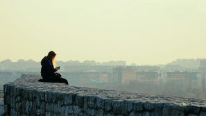Una ragazza seduta su un muretto che si affaccia sulla città al tramonto mentre guarda il suo telefono in solitudine