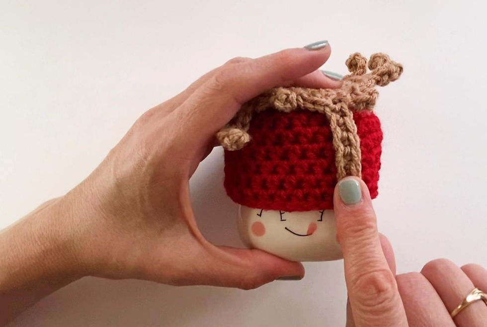 Hands gluing crocheted "caramel" onto a crocheted apple hat worn by a marshmallow mug.