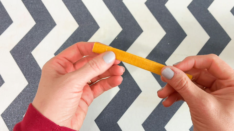 Hands holding a yellow strip against a gray and white chevron-patterned background. The person wears a red sleeve and has light nail polish.
