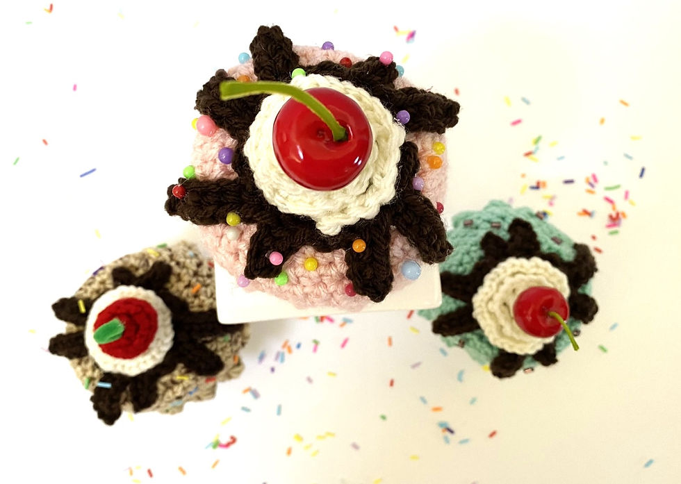 A view of three crocheted marshmallow mug hats that look like ice cream with whipped cream, chocolate topping nad sprinkles.