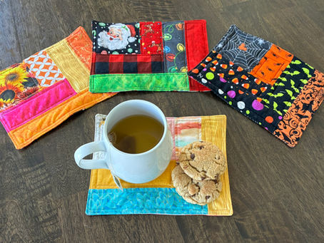 Colorful quilted mug rugs on a wooden table with tea in a white mug and cookies. Festive patterns include holiday and Halloween themes.