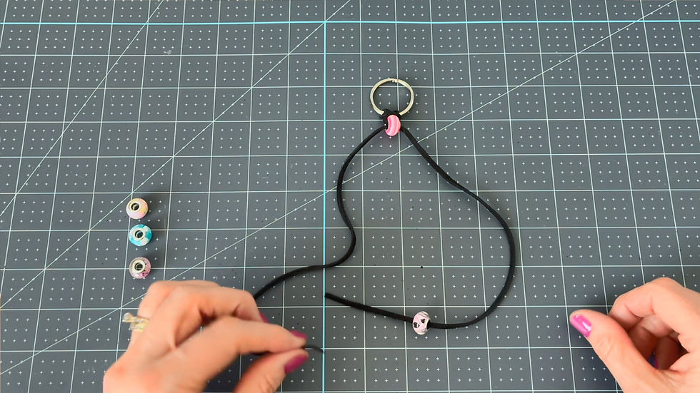 Hands string beads onto black cord on a grid-patterned mat. A ring binder clip and colorful beads are visible. Bright pink nail polish stands out.