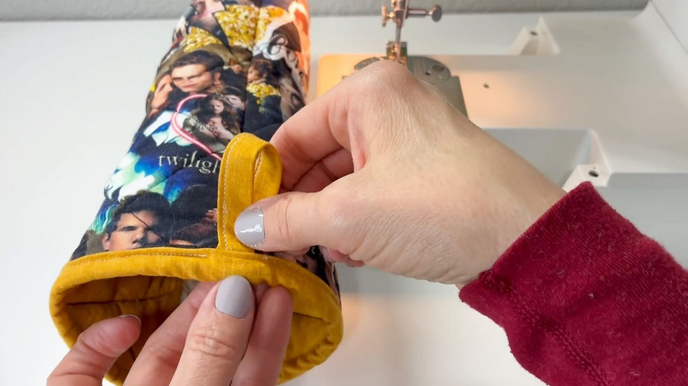 Hands in red sleeves hold a colorful oven mitt with images and "Twilight" text by a sewing machine, highlighting a yellow loop.