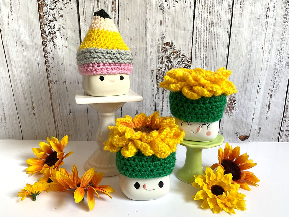 Three marshmallow mugs wearing crocheted hats that look like sunflowers and a pencil.