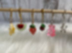 Colorful fruit and candy stitch markers for crochet hanging on a wooden rod: banana, watermelon, strawberry, cherry, gummy bear, and balloon dog. White fluffy background.