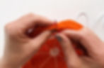 Hands using a darning needle to weave elastic through the last row of a round crocheted potholder that looks like a slice of an orange.