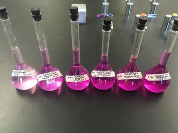 A series of 6 volumetric flasks hold pink solutions of varying concentrations. The flasks are lined up on a black lab bench.