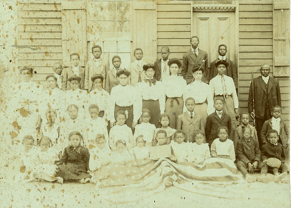 A historic sepia-toned group photograph, with age spots and staining, of Black students and teachers standing and seated in rows outside a wooden schoolhouse. Some children in the front row are seated on an American flag laid on the ground. A man on the right holds a rifle.