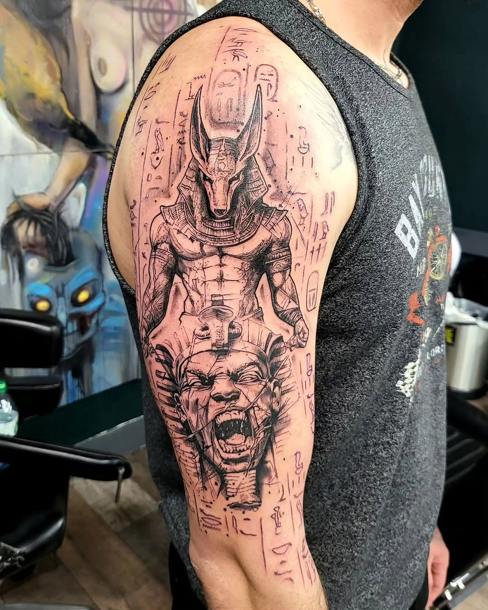 Arm showing a black ink Anubis tattoo and roaring face tattoo in a tattoo shop, with stencil marks and graffiti-like wall art.