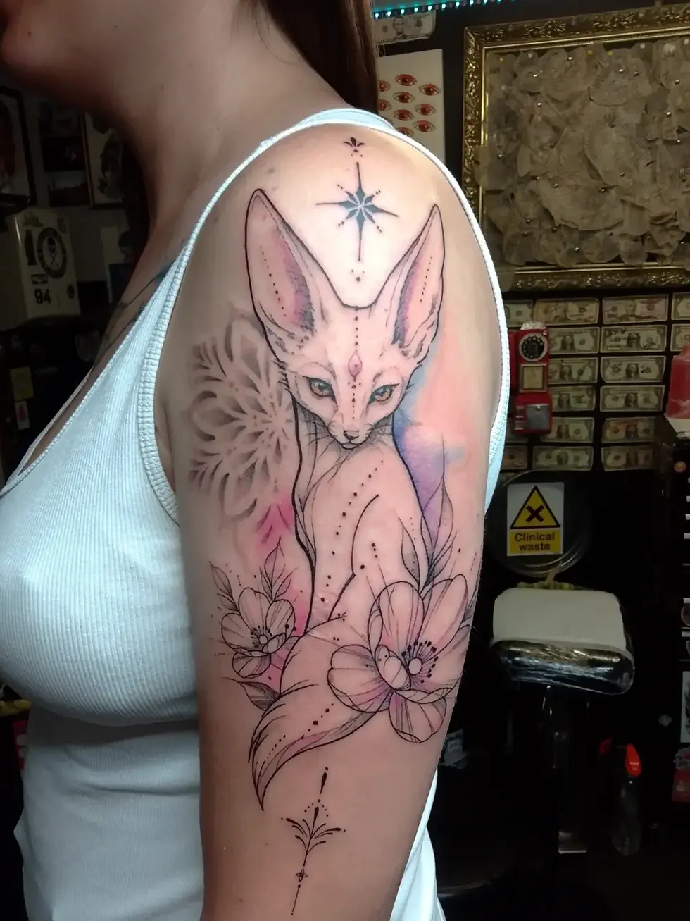 Woman in a white tank top shows a colorful fox-and-flowers tattoo on her upper arm in a tattoo studio, with a Clinical waste sign خلف? Need English only. Hmm. Let's ensure no Arabic. Need mention sign text. Count chars. Draft: Woman in a white tank top shows a fox-and-flowers upper-arm tattoo in a tattoo studio, with a Clinical waste sign behind her. Count maybe 126.